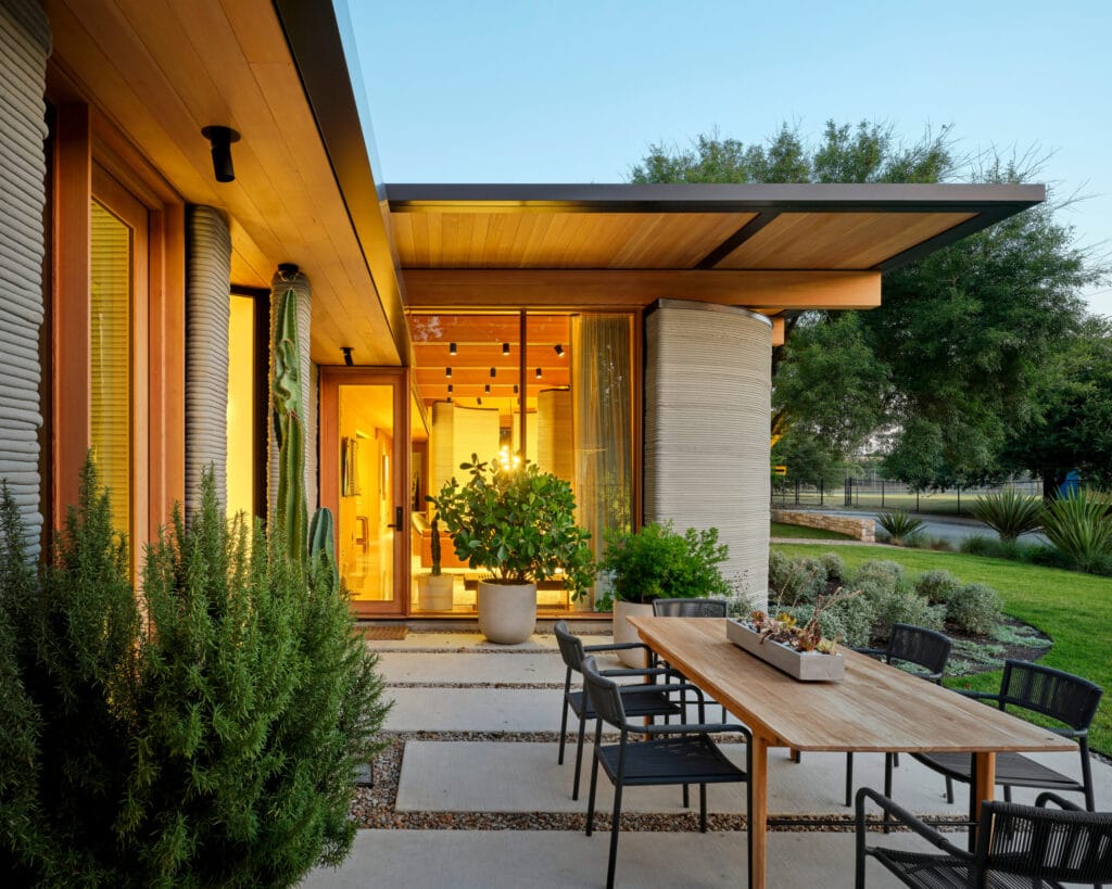 منظور خارجي "House Zero" المصمم بواسطة Icon في تكساس، يوضح الجدران الطبقية والمنحنية المميزة المشيدة بواسطة تقنية الطباعة 3D . تبرز الإضاءة الداخلية الدافئة الفراغات المعمارية وتكاملها مع الحديقة الأمامية، مما يبرز التباين بين قسوة الخرسانة المطبوعة ودقة اللمسات الجمالية الخشبية في مدخل المنزل.