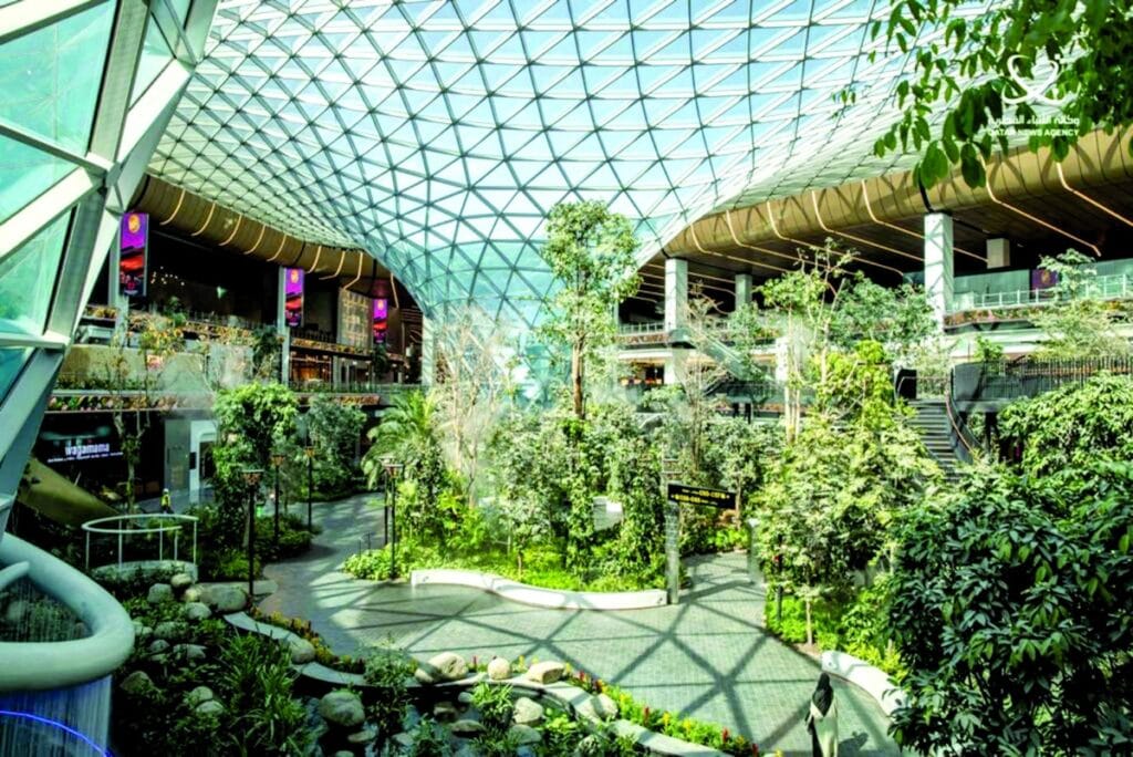 لقطة بانورامية لمنطقة أورتشارد (The Orchard) في مطار حمد الدولي بالدوحة، تظهر الحديقة الاستوائية الداخلية الممتدة على مساحة 6,000 متر مربع تحت سقف زجاجي انسيابي ضخم بنظام النمذجة البارامترية، مع ممرات للمشاة محاطة بأشجار شاهقة وتفاصيل معمارية بارزة