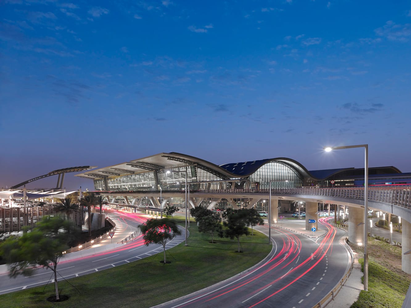 لقطة ليلية لواجهة مطار حمد الدولي، تُظهر التموجات السقفية المستلهمة من حركة الأمواج والكثبان الرملية، مع إبراز اتصال المبنى بشبكة الطرق الانسيابية والجسور، في مشهد يجمع بين الإضاءة الحيوية وتكامل النظام الإنشائي مع البيئة المحيطة