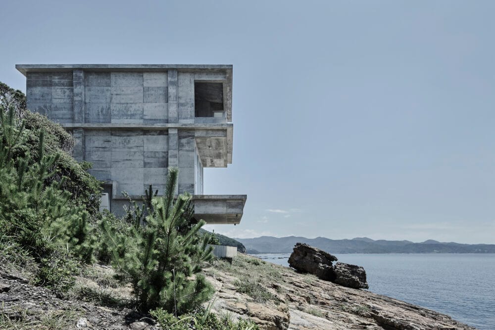 منظور جانبي لمنزل 'Yasun House' في اليابان، يبرز التضاد الحاد بين كتلته الخرسانية العارية (الخام) وبين شفافية الواجهة الزجاجية الغاطسة. ويُظهر التصميم الانزلاق الكتلي للطوابق الذي يخلق شرفات مظللة تؤطر إطلالة بانورامية على المحيط من فوق منحدر جبلي وعر.