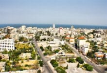 إطلالة نهارية علوية على مدينة غزة تبرز المناطق الخضراء والمساحات المفتوحة وسط النسيج العمراني الحديث، مع ظهور شوارع رئيسية منظمة ومباني بيضاء تميز حي الرمال، وينتهي الأفق بالخط الأزرق الصافي لساحل البحر