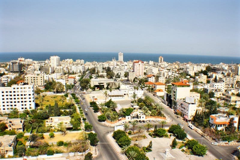 إطلالة نهارية علوية على مدينة غزة تبرز المناطق الخضراء والمساحات المفتوحة وسط النسيج العمراني الحديث، مع ظهور شوارع رئيسية منظمة ومباني بيضاء تميز حي الرمال، وينتهي الأفق بالخط الأزرق الصافي لساحل البحر