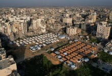 لقطة جوية لحي سكني في مدينة غزة المدمرة، تظهر مخيماً منظماً لوحدات إيواء جاهزة مستطيلة ودائرية تم زرعها ككتلة عازلة وسط نسيج حضري مدمر بالكامل.