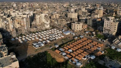 لقطة جوية لحي سكني في مدينة غزة المدمرة، تظهر مخيماً منظماً لوحدات إيواء جاهزة مستطيلة ودائرية تم زرعها ككتلة عازلة وسط نسيج حضري مدمر بالكامل.