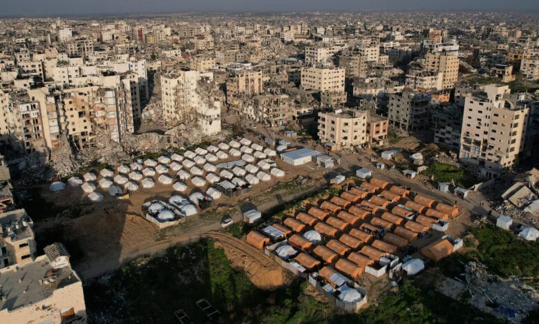 لقطة جوية لحي سكني في مدينة غزة المدمرة، تظهر مخيماً منظماً لوحدات إيواء جاهزة مستطيلة ودائرية تم زرعها ككتلة عازلة وسط نسيج حضري مدمر بالكامل.