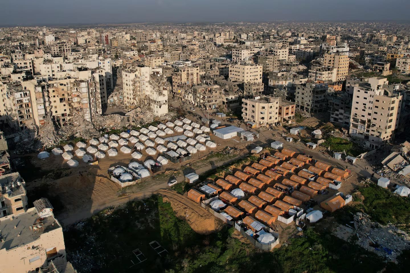 لقطة جوية لحي سكني في مدينة غزة المدمرة، تظهر مخيماً منظماً لوحدات إيواء جاهزة مستطيلة ودائرية تم زرعها ككتلة عازلة وسط نسيج حضري مدمر بالكامل.
