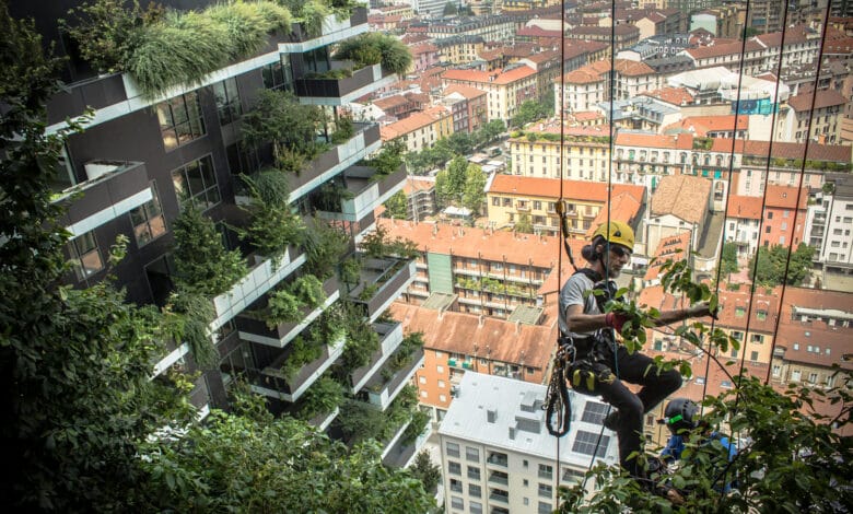 لقطة مقربة تُظهر فريق صيانة متخصص يمارس أعمال التقليم والرعاية للنباتات الكثيفة على شرفات مبنى Bosco Verticale في ميلانو، مما يجسد الجانب العملي والمهني المطلوب للحفاظ على الاستدامة العمرانية في الغابات العمودية.