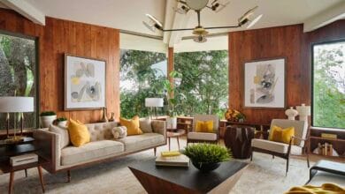 فراغ معيشة يجسد طراز Mid-Century Modern بكسوة جدران خشبية دافئة، أثاث بأرجل مدببة (Tapered Legs)، وثريا أيقونية، تطل على مساحات خضراء عبر نوافذ واسعة.