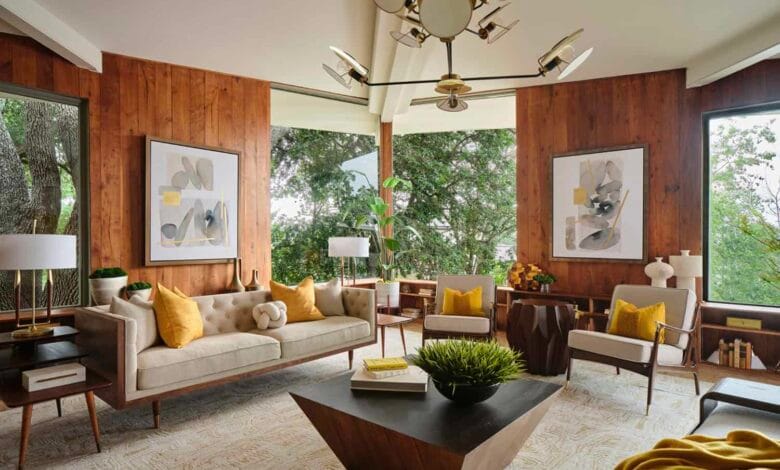 فراغ معيشة يجسد طراز Mid-Century Modern بكسوة جدران خشبية دافئة، أثاث بأرجل مدببة (Tapered Legs)، وثريا أيقونية، تطل على مساحات خضراء عبر نوافذ واسعة.