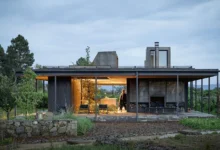 لقطة لمنزل 'Rain Harvest Home' في المكسيك، تُظهر هيكلاً معمارياً بسيطاً من الفولاذ والخشب يتميز بسطح مستوٍ مصمم لجمع مياه الأمطار، مع فضاءات داخلية مفتوحة تتداخل مع الطبيعة المحيطة وإضاءة دافئة تبرز شفافية التصميم في الغسق