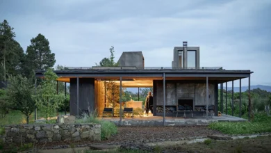 لقطة لمنزل 'Rain Harvest Home' في المكسيك، تُظهر هيكلاً معمارياً بسيطاً من الفولاذ والخشب يتميز بسطح مستوٍ مصمم لجمع مياه الأمطار، مع فضاءات داخلية مفتوحة تتداخل مع الطبيعة المحيطة وإضاءة دافئة تبرز شفافية التصميم في الغسق