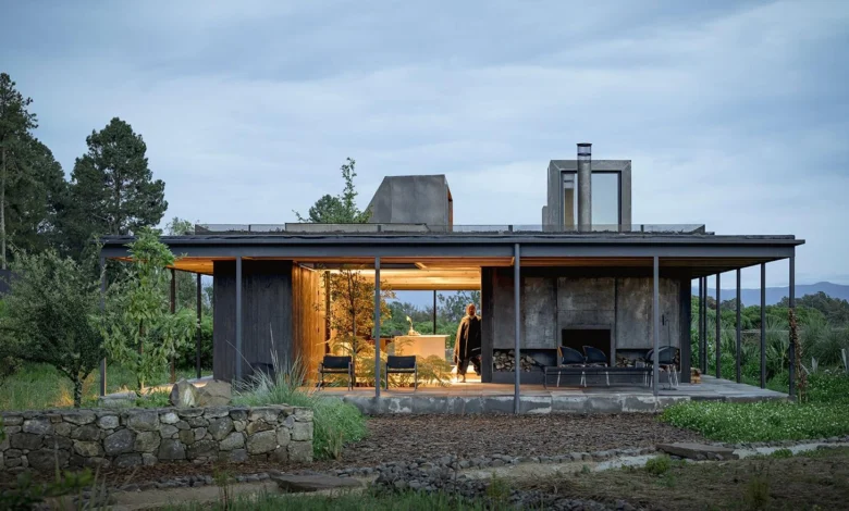 لقطة لمنزل 'Rain Harvest Home' في المكسيك، تُظهر هيكلاً معمارياً بسيطاً من الفولاذ والخشب يتميز بسطح مستوٍ مصمم لجمع مياه الأمطار، مع فضاءات داخلية مفتوحة تتداخل مع الطبيعة المحيطة وإضاءة دافئة تبرز شفافية التصميم في الغسق