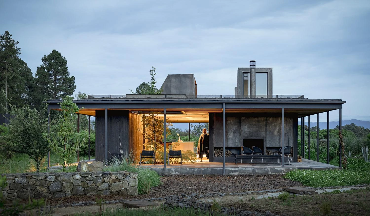 لقطة لمنزل 'Rain Harvest Home' في المكسيك، تُظهر هيكلاً معمارياً بسيطاً من الفولاذ والخشب يتميز بسطح مستوٍ مصمم لجمع مياه الأمطار، مع فضاءات داخلية مفتوحة تتداخل مع الطبيعة المحيطة وإضاءة دافئة تبرز شفافية التصميم في الغسق