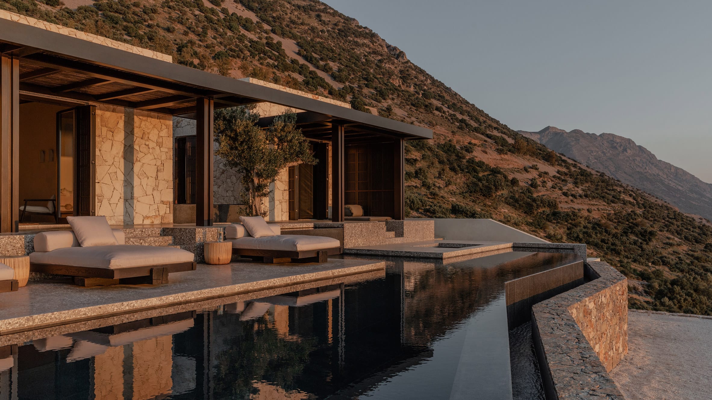 منظور معماري لمنزل 'Hill House' في جزيرة كريت، اليونان، يبرز تصميماً غاطساً جزئياً في التلة يتألف من كتل خرسانية أفقية متدرجة وشرفات واسعة تطل مباشرة على البحر، مع سقف أخضر يدمج الهيكل بالكامل في الطبيعة الجبلية المحيطة.