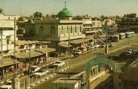 صورة أرشيفية ملونة لوسط مدينة غزة تبرز عمارة السبعينيات؛ حيث يظهر مبنى تاريخي تعلوه قبة خضراء مميزة، مع حركة السيارات والحافلات القديمة في الشارع الرئيسي الذي يربط أحياء المدينة، مما يعكس النسيج العمراني والنشاط التجاري في تلك الحقبة