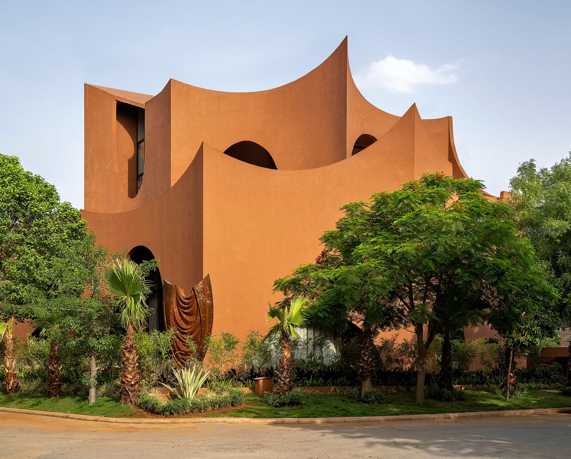 واجهة منزل 'ميراي' (Mirai House of Arches) المصبوغة باللون الطيني المحروق، تبرز سلسلة من الأقواس والمنحنيات النحتية الضخمة المتداخلة، والمصممة خصيصاً لخلق مناطق تظليل طبيعية وتهوية متقاطعة في مواجهة المناخ الصحراوي القاسي، مع تكامل بستان من الأشجار الخضراء في المقدمة