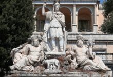 لقطة خارجية توضح نافورة "Dea Roma" (إلهة روما) في ساحة ديل بوبولو، حيث يظهر تمثال مركزي لروما محاطاً بتماثيل تمثل نهري التيبر والأنياني، مع خلفية معمارية كلاسيكية تبرز العقود والزخارف الرومانية التاريخية.