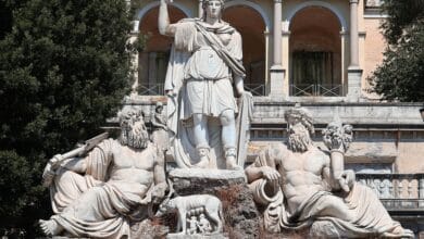 لقطة خارجية توضح نافورة "Dea Roma" (إلهة روما) في ساحة ديل بوبولو، حيث يظهر تمثال مركزي لروما محاطاً بتماثيل تمثل نهري التيبر والأنياني، مع خلفية معمارية كلاسيكية تبرز العقود والزخارف الرومانية التاريخية.