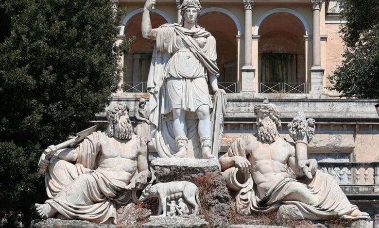 لقطة خارجية توضح نافورة "Dea Roma" (إلهة روما) في ساحة ديل بوبولو، حيث يظهر تمثال مركزي لروما محاطاً بتماثيل تمثل نهري التيبر والأنياني، مع خلفية معمارية كلاسيكية تبرز العقود والزخارف الرومانية التاريخية.