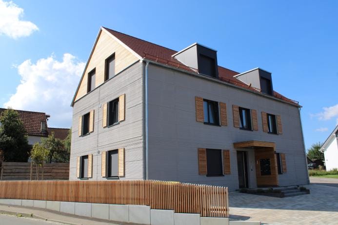 منظور خارجي لمبنى سكني من ثلاثة طوابق في Wallenhausen بألمانيا، تبرز الصورة الجدران الرمادية ذات الطبقات الإنشائية والأسقف المائلة القرميدية، مما يجسد تكامل حلول الاسكان المستدامة مع الهوية المعمارية الريفية للمنطقة.