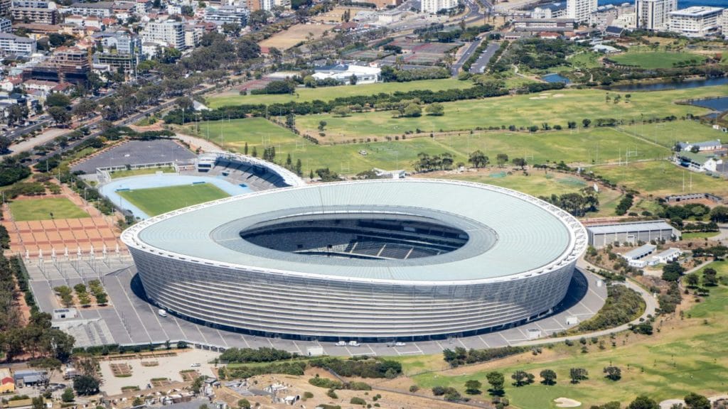 منظور جوي لملعب كيب تاون 'Cape Town Stadium' ذو الواجهة البيضاء المتموجة، يوضح تموضعه على حافة المحيط بعيداً عن مركز المدينة النابض، مما يبرز عقم التصميم المعتمد على 'الأيقونة' التي تتحول لفراغ ميت يمتص ميزانيات الصيانة بعد البطولة.