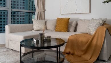 أريكة زاوية مودرن مريحة بلون بيج فاتح، مزينة بغطاء خردلي وطاولة قهوة زجاجية دائرية.