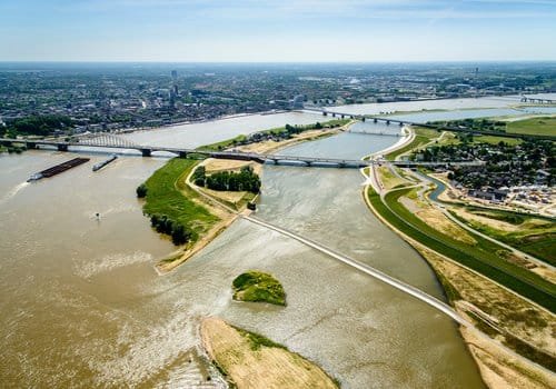منظر جوي بانورامي لمدينة نيميغن الهولندية، يُظهر التطبيق العملي لمفهوم "Room for the River"؛ حيث يبدو نهر "فال" (Waal) وقد تم منح المياه مساحة إضافية للتمدد من خلال حفر قناة جانبية واسعة، مما خلق جزيرة حضرية جديدة مدمجة بالكامل مع النسيج العمراني عبر جسور حديثة وتخطيط لاندسكيب مرن يعزز التكيف مع الفيضانات